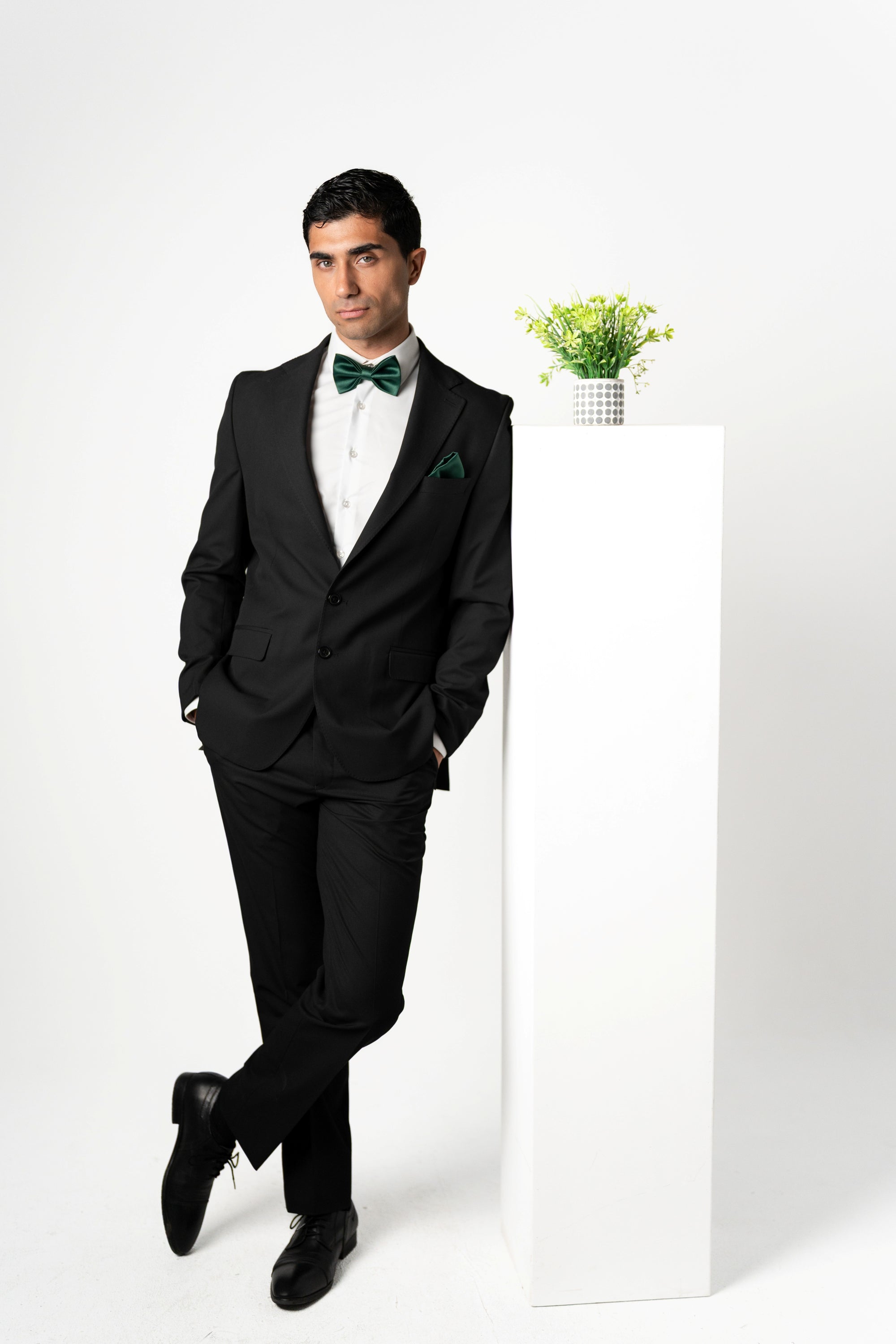 Man in a black suit with a green bow tie standing against a white background