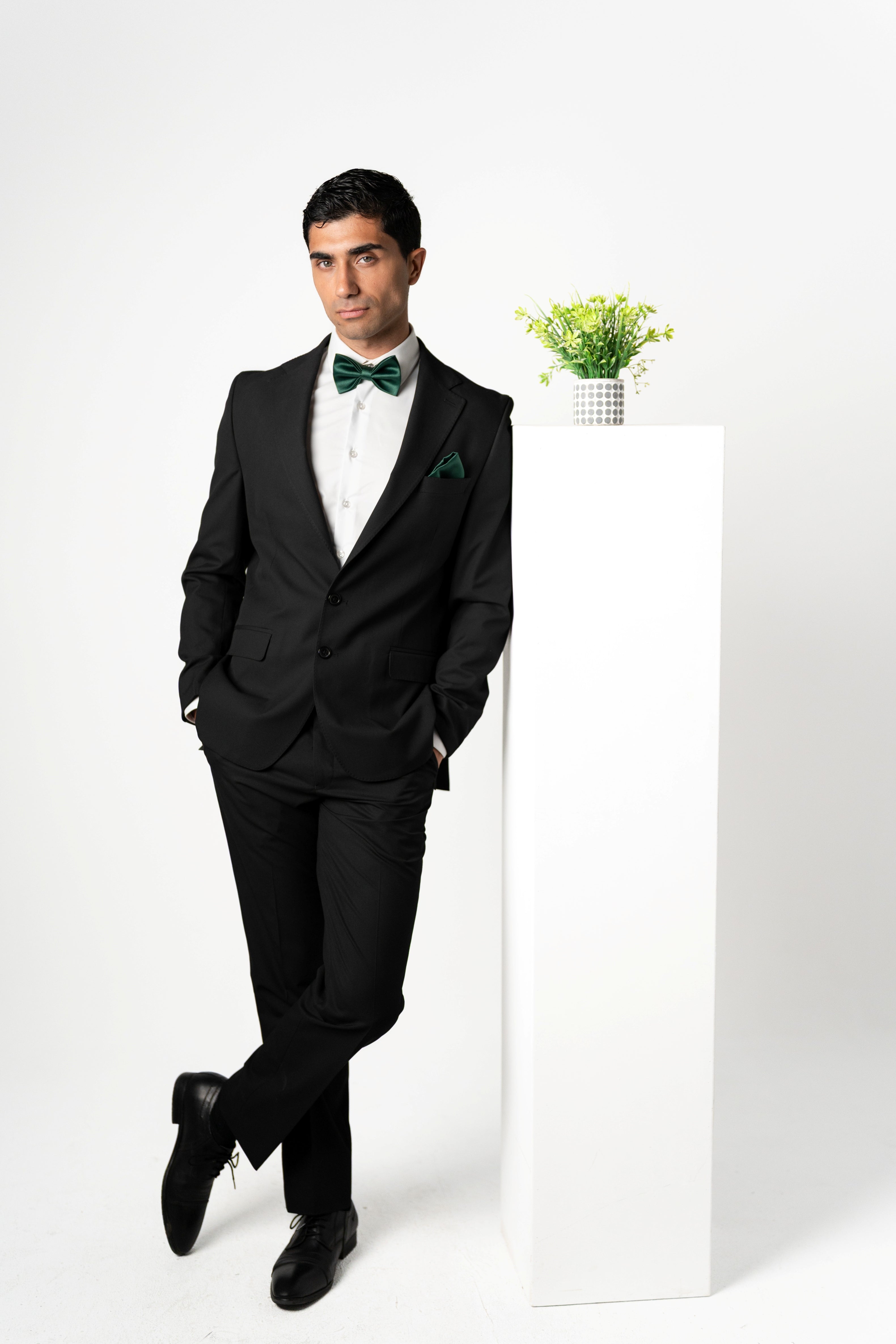 Man in a black suit with a green bow tie standing against a white background