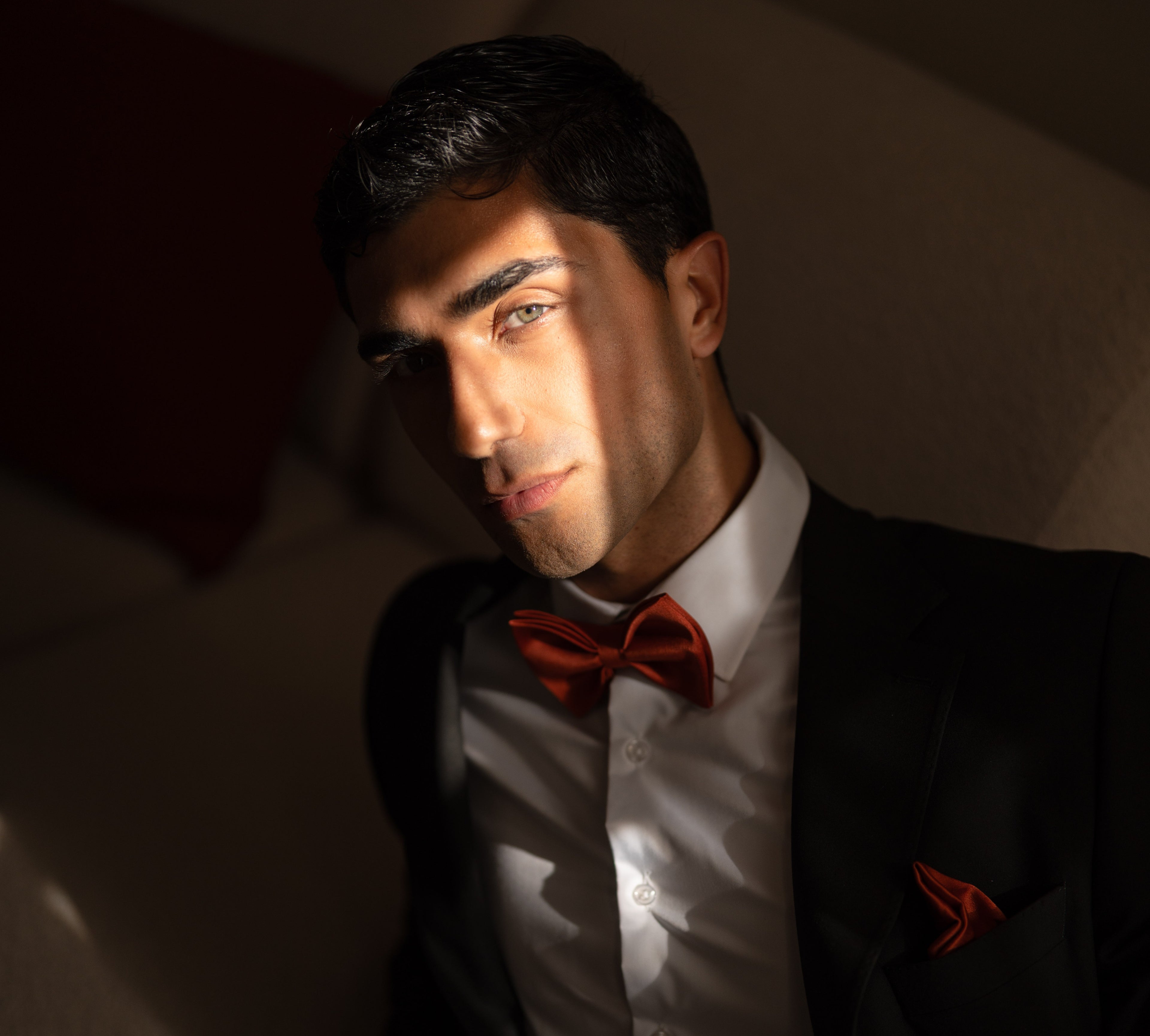 Man wearing a black suit with a red bow tie and pocket square against a dark background